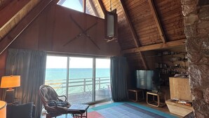 Ein gemütliches Zimmer mit schrägen Wänden, einem großen Fenster mit Meerblick, einem bequemen Sessel, einer Couch, einem Flachbildfernseher und einer Steinwand.