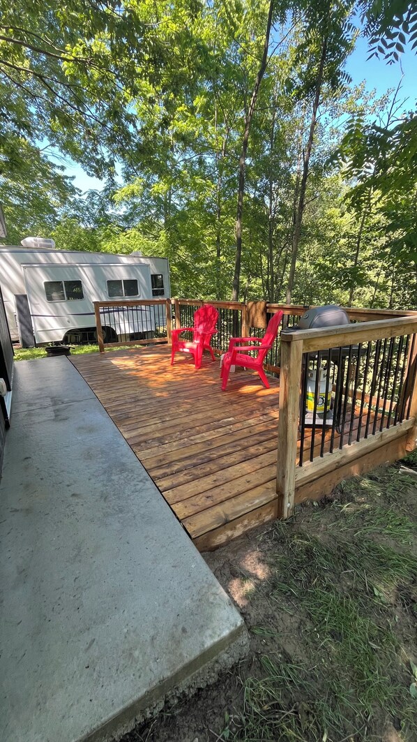 A wooden deck with a concrete pathway, surrounded by trees and a white RV.