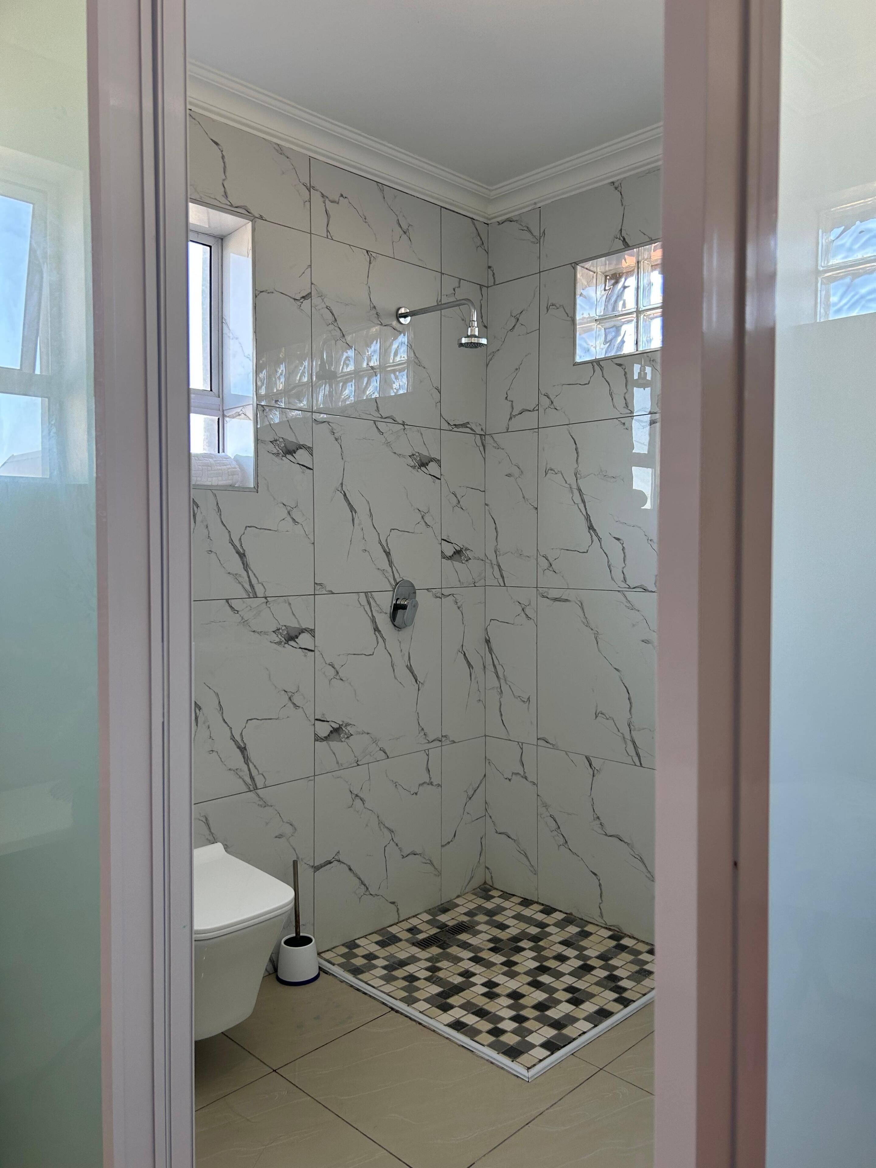 Et badeværelse med toilet, brusebad med marmorvæg og et skakternet gulv.