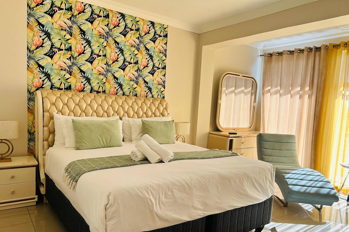 Une chambre d’hôtel avec un grand lit, une table de chevet, une chaise, un miroir et une fenêtre avec des rideaux.