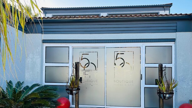 Entrada de hotel con un diseño moderno, con dos grandes puertas de vidrio que muestran el número '5' y la inscripción 'FIFTY Boutique Hotel & Spa'.