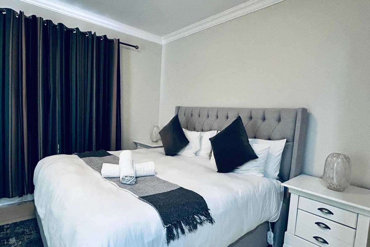 Une chambre d’hôtel avec un grand lit, une table de chevet, une commode et une fenêtre avec des rideaux.