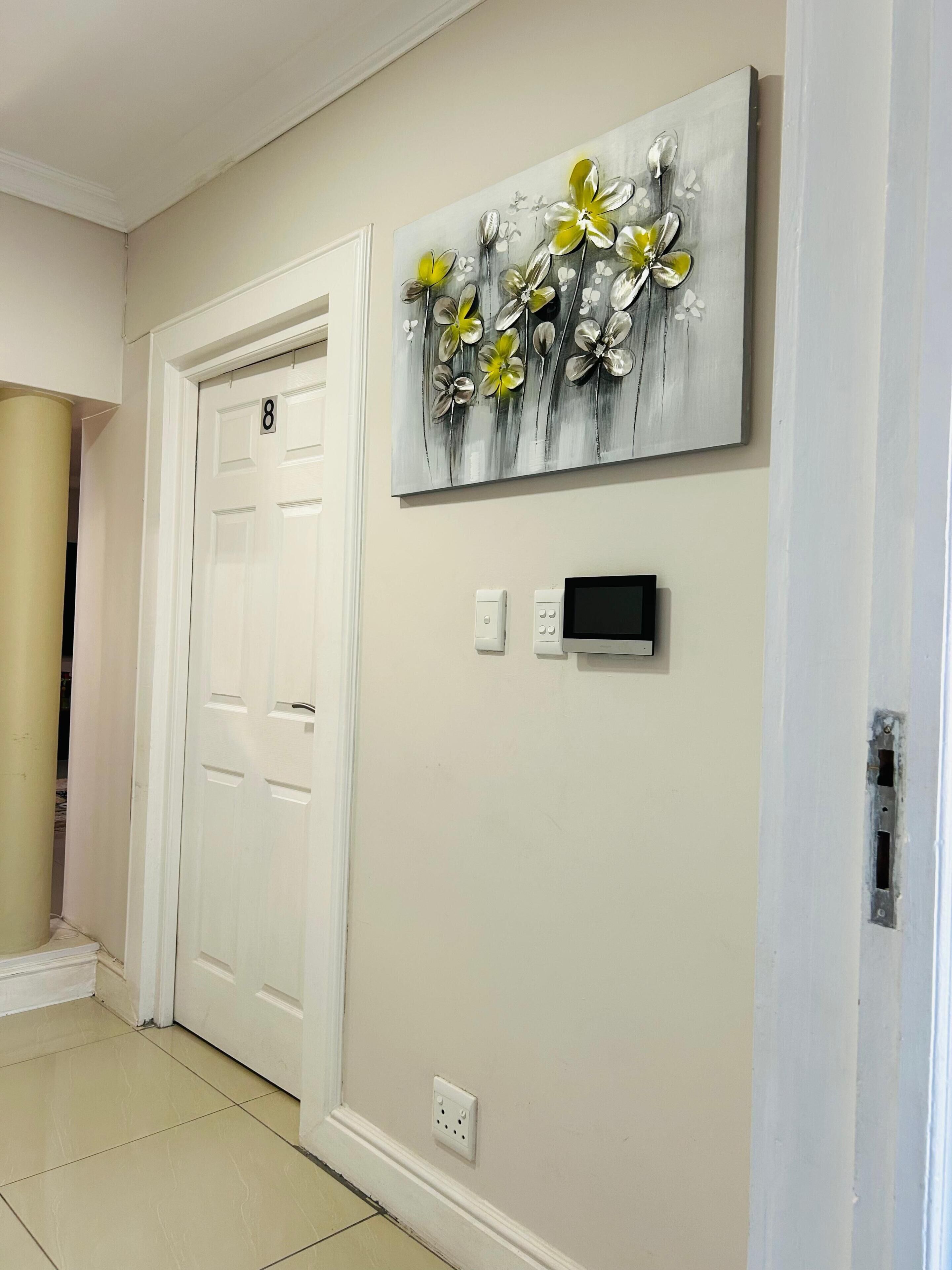 A hallway with a white door marked '8', a wall-mounted artwork of flowers, a thermostat, and a power outlet.