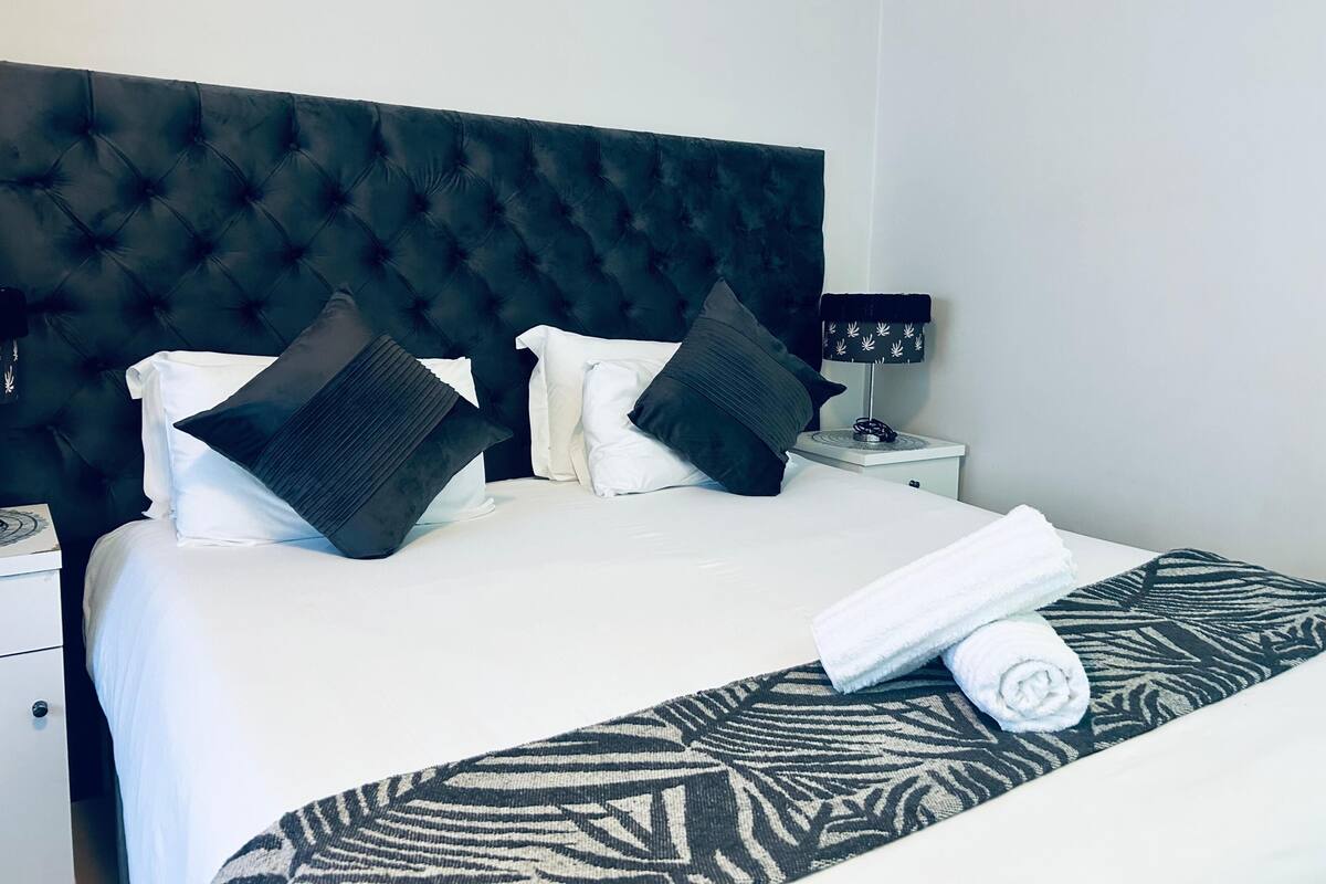 Une chambre d’hôtel avec une tête de lit capitonnée, un lit avec des oreillers blancs et foncés, une table de chevet avec une lampe et une couvre-lit à motifs.