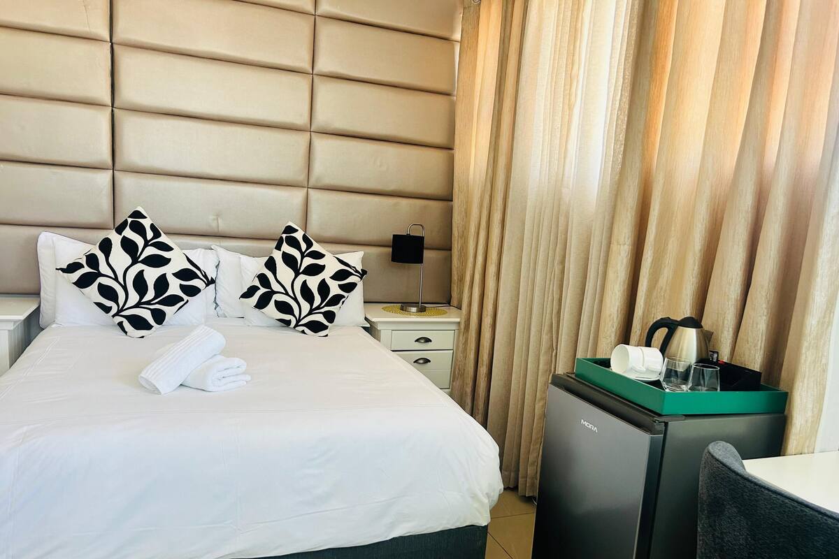 Une chambre d’hôtel comprenant un lit, deux coussins décoratifs, une table de chevet avec une lampe, un petit réfrigérateur et une chaise.