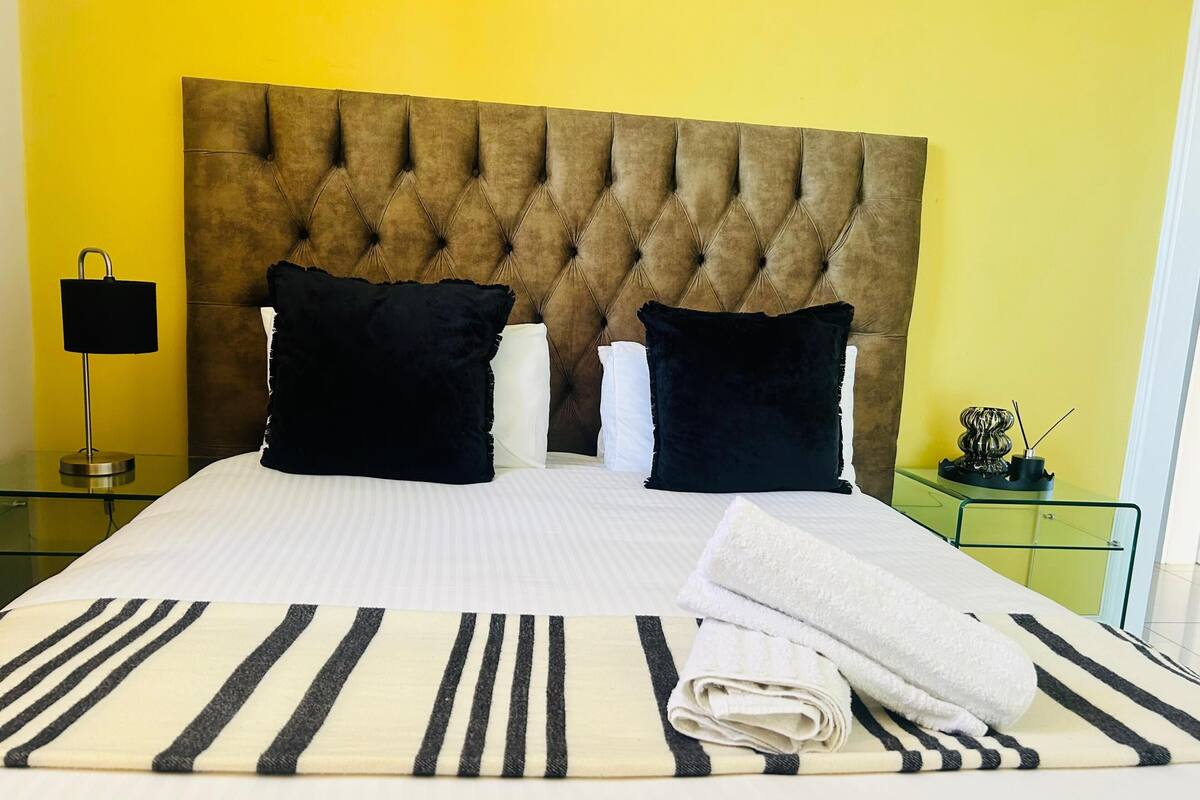 Une chambre d’hôtel avec une tête de lit capitonnée, une couvre-lit à rayures, des oreillers noirs et blancs, une table de chevet en verre avec une lampe, et une table de nuit verte agrémentée d’un objet décoratif.