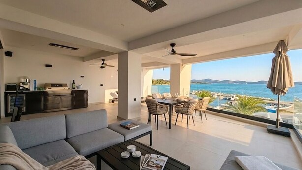Una sala moderna con sofá, mesa de centro y zona de comedor. Cuenta con un amplio ventanal con vista al mar y a las montañas.