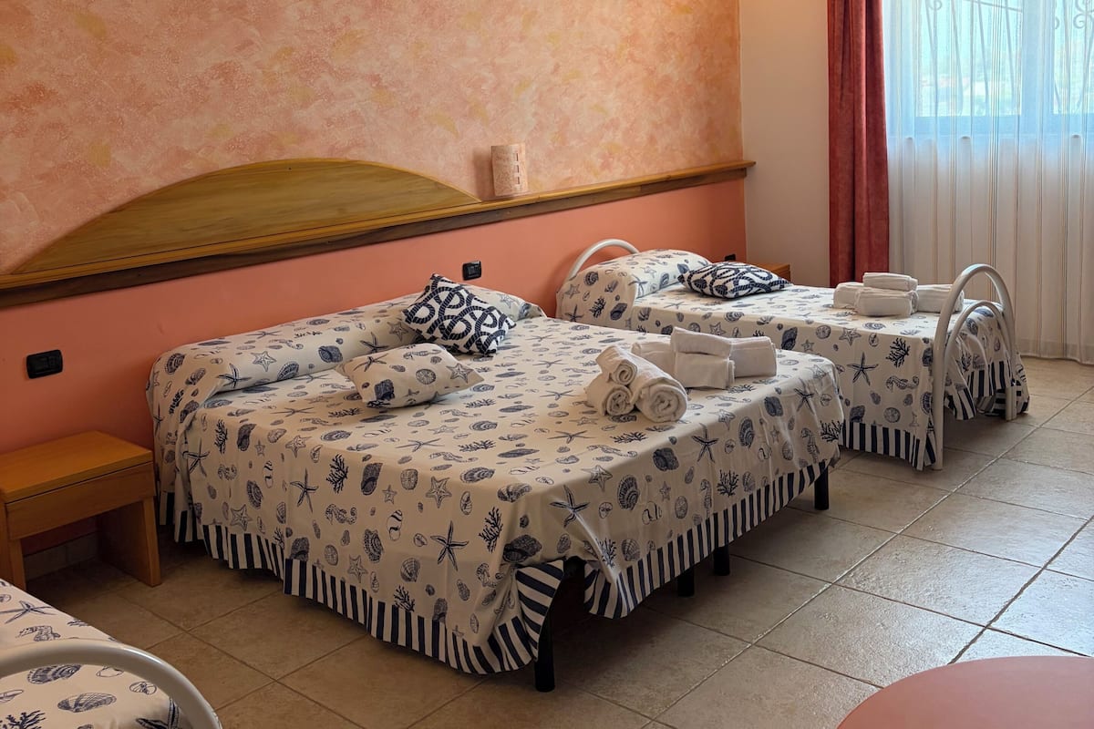 Habitació d'hotel amb tres llits, una taula petita, un capçal de llit i una finestra amb cortines.