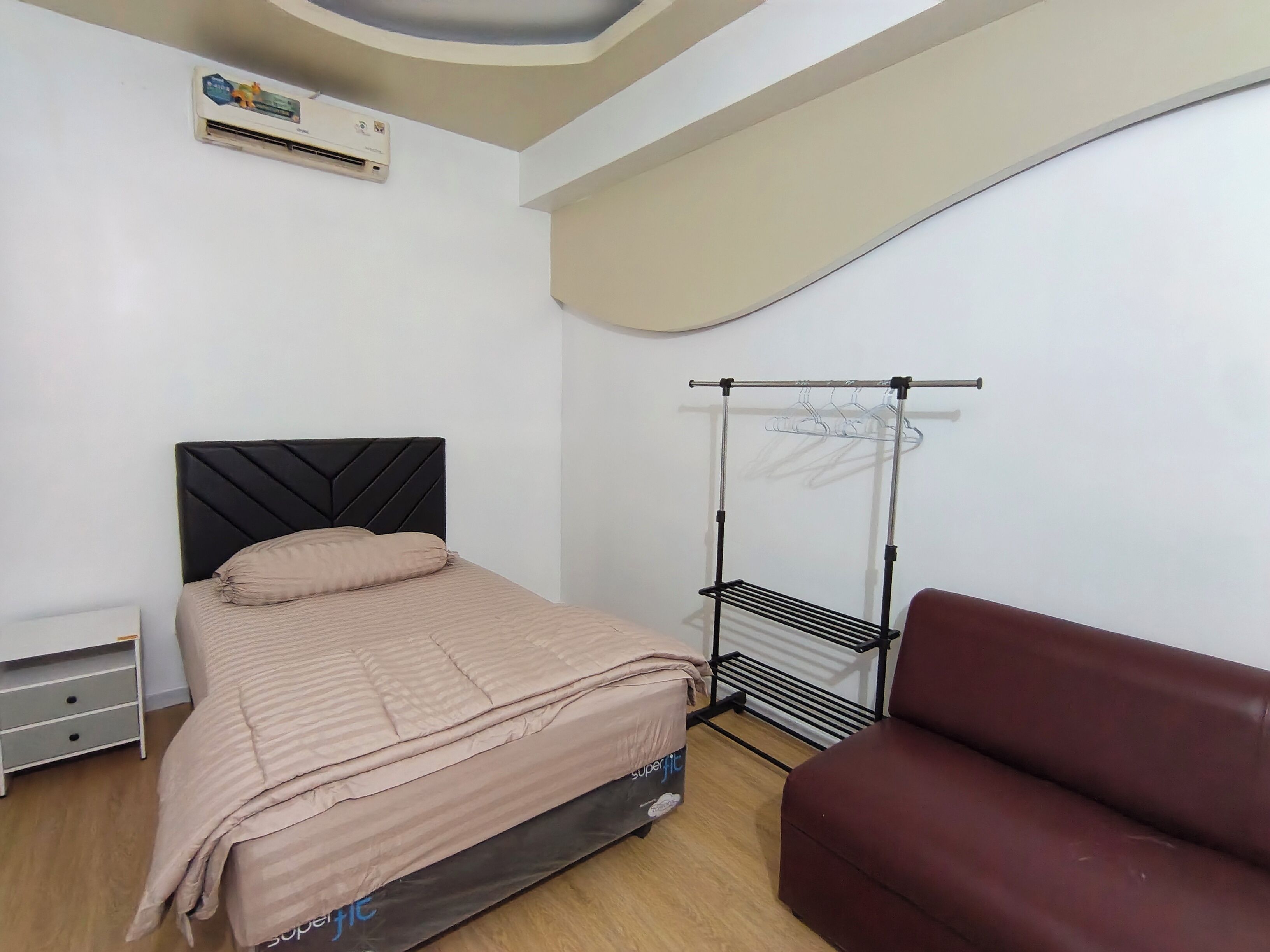 A bedroom with a bed, a red couch, a wall-mounted air conditioner, and a drying rack.