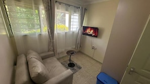 A small living area with a sofa, a TV mounted on the wall, a small table, and a window with curtains. - Golden Studio (Nairobi)
