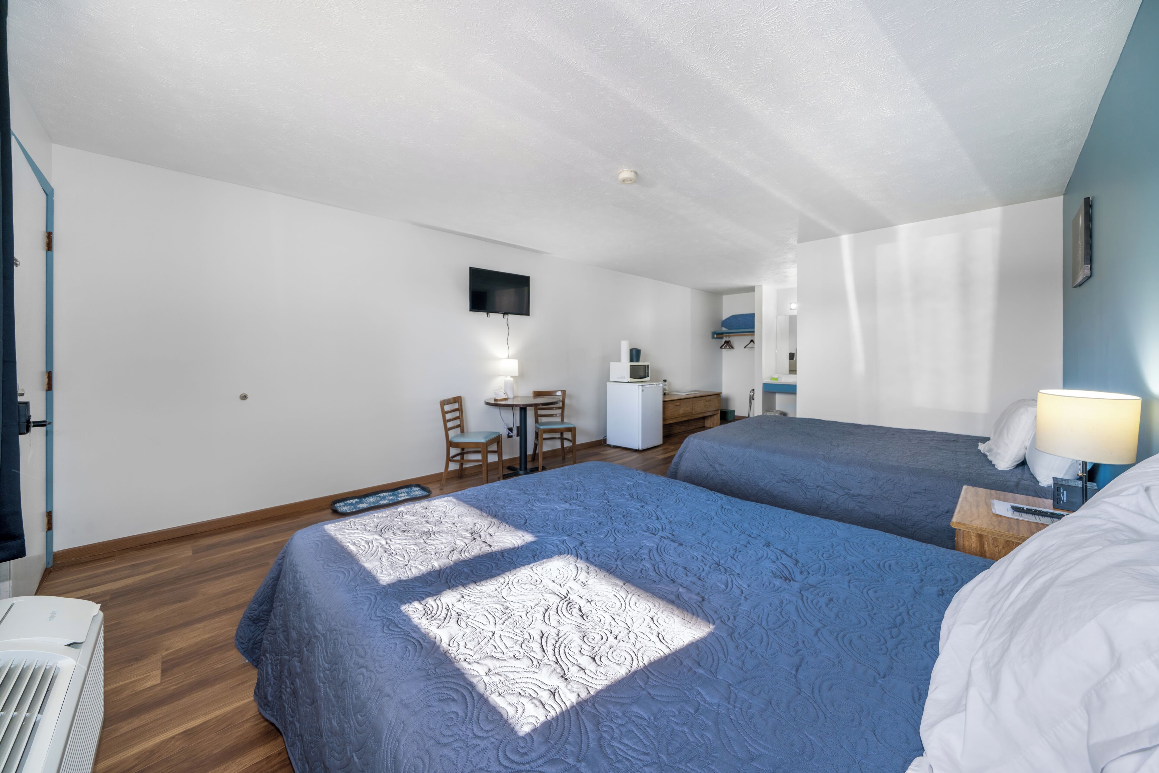 Habitación de hotel con dos camas, un escritorio, una silla, un televisor montado en la pared y una pequeña zona de kitchenette.