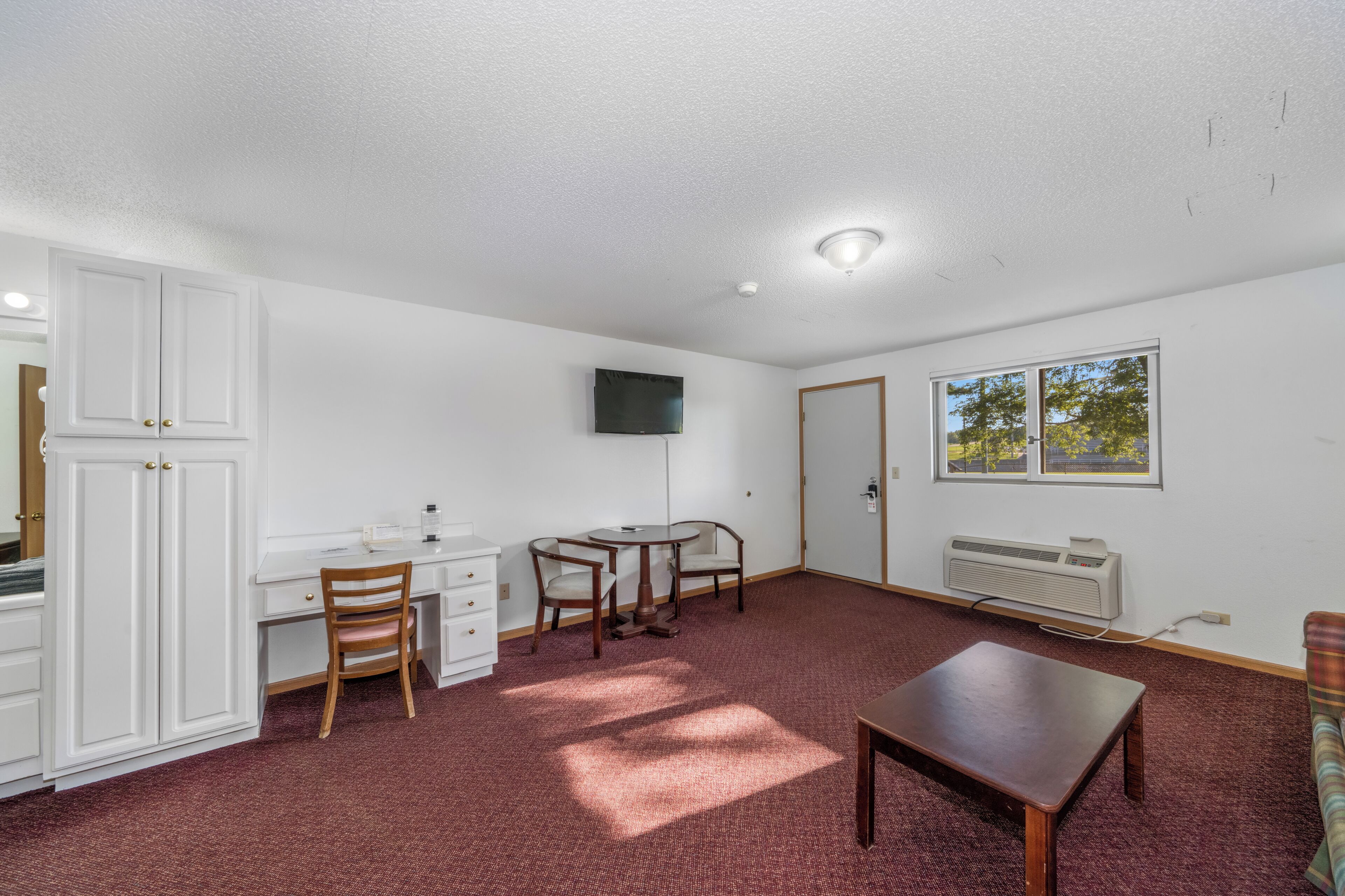 A room with a desk, two chairs, a television mounted on the wall, a radiator, and a window with a view of trees.