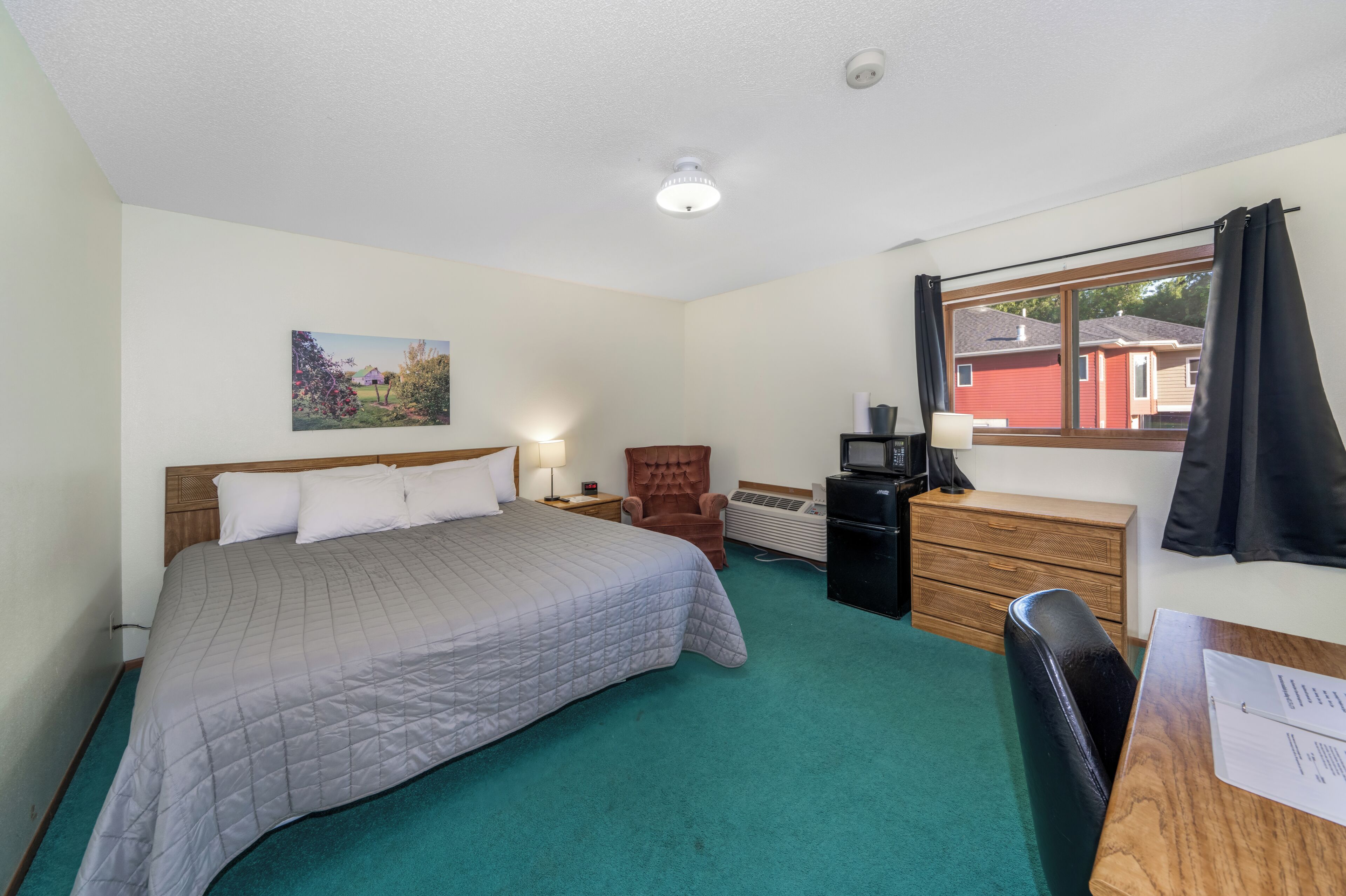 A hotel room with a bed, a desk, a chair, a heater, a window with curtains, and a painting on the wall.