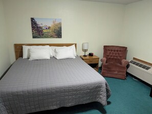 A neatly made bed with a quilted cover, a brown upholstered armchair, a bedside table with a lamp, and a wall-mounted picture. - Auburn Inn South (Auburn)