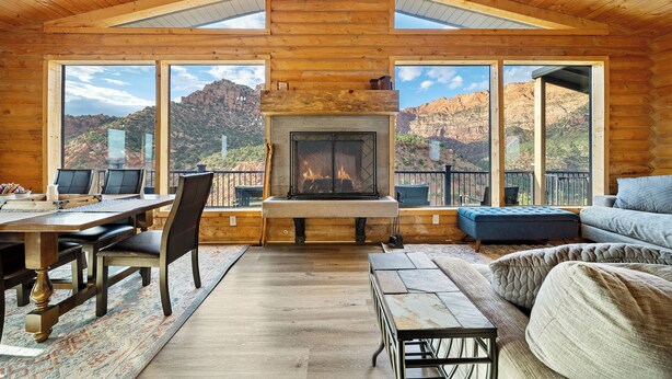 A log cabin interior with a fireplace, large windows, a dining area, and a seating area with a view of the mountains. - Elevation40Zion (Apple Valley)