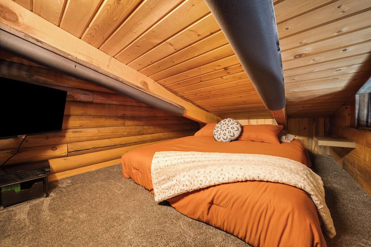 Interior de uma cabana de madeira com uma cama, uma televisão e um respiradouro no teto.