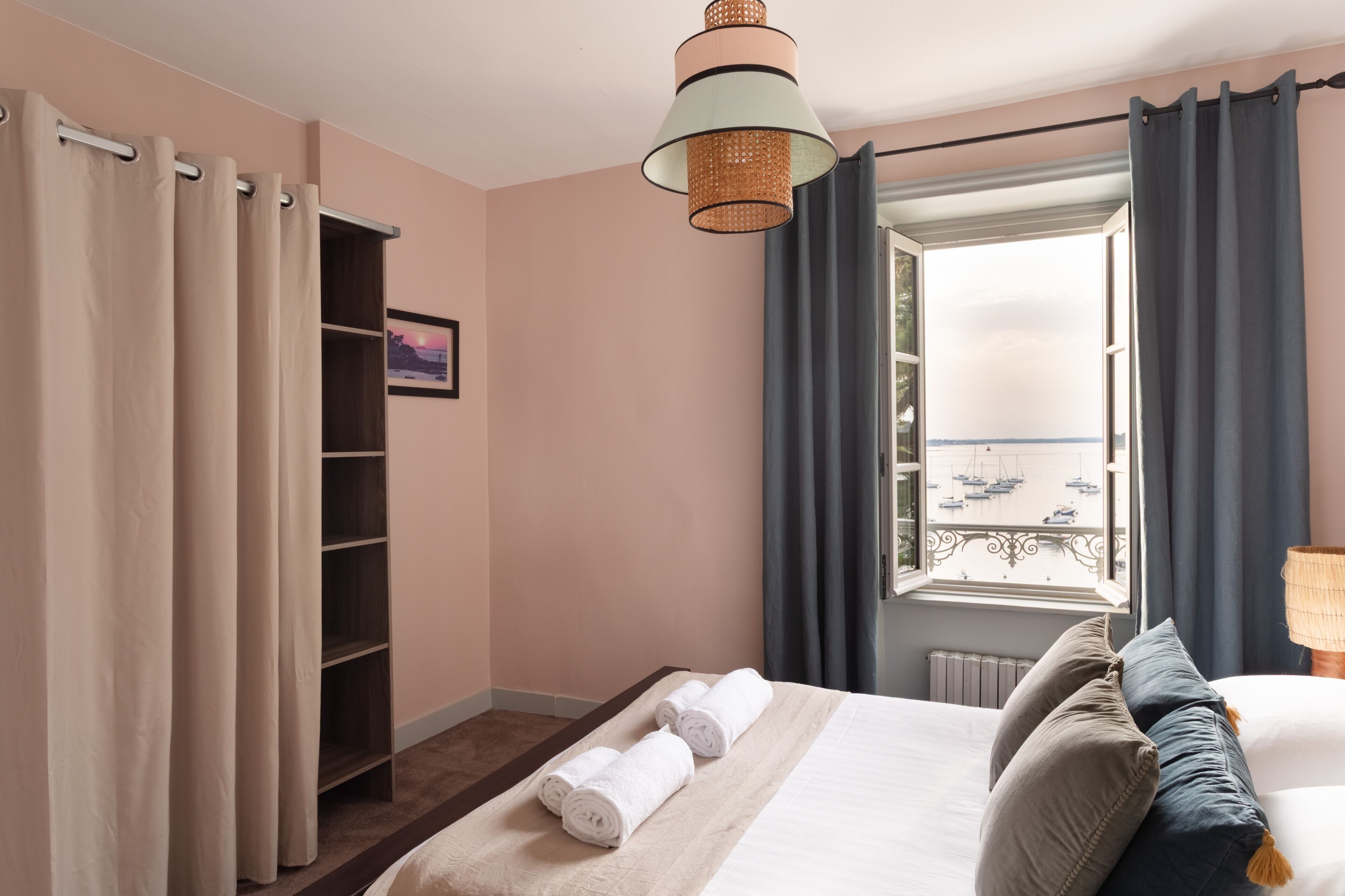 A hotel room with a bed, a window with a view of boats, a shelf, and a lamp.