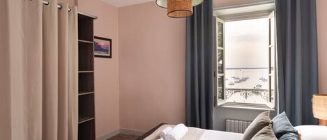 A hotel room with a bed, a window with a view of boats, a shelf, and a lamp.