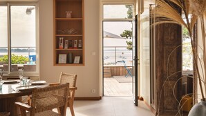 Ein Essbereich mit Tisch für vier Personen, Korbstühle, einem Holzregal mit Dekoration und Blick auf das Meer durch eine Glastür.