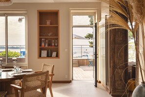 Un coin repas avec une table dressĂ©e pour quatre personnes, des chaises en osier, une bibliothĂšque en bois agrĂ©mentĂ©e dâobjets dĂ©coratifs et une vue sur la mer par une porte vitrĂ©e.