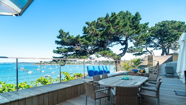 Eine Holzterrasse mit Tisch und Stühlen, mit Blick auf ein Gewässer und Boote.