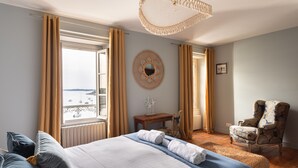 Ein Schlafzimmer mit Bett, Stuhl, Tisch, Spiegel und Blick auf das Meer.