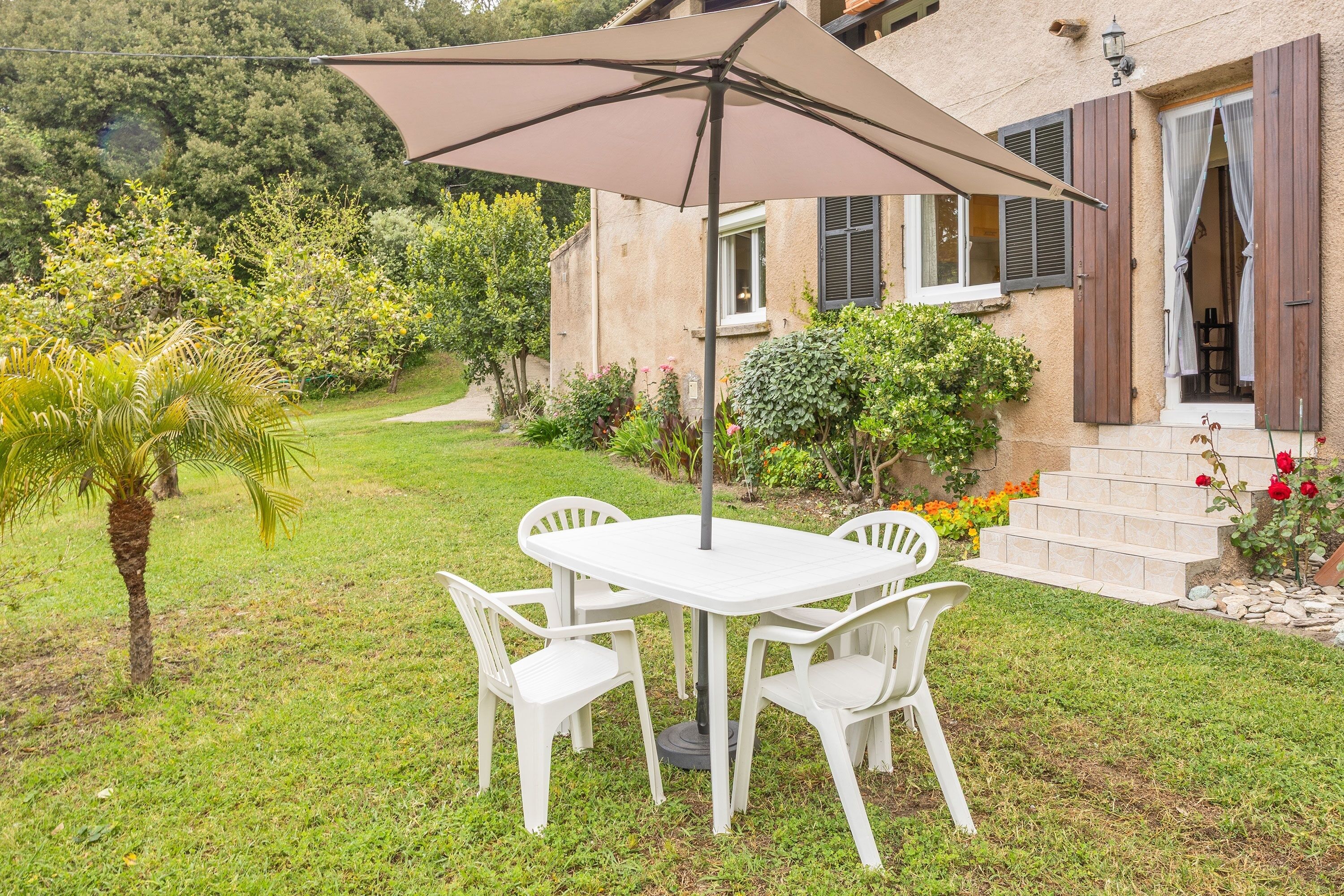 Een terras met een tafel, stoelen, een grote parasol en een huis met luiken.