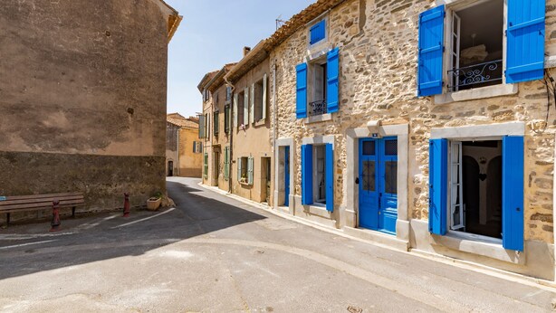 Una calle estrecha con edificios de piedra, algunos con contraventanas azules, y un banco a la izquierda.