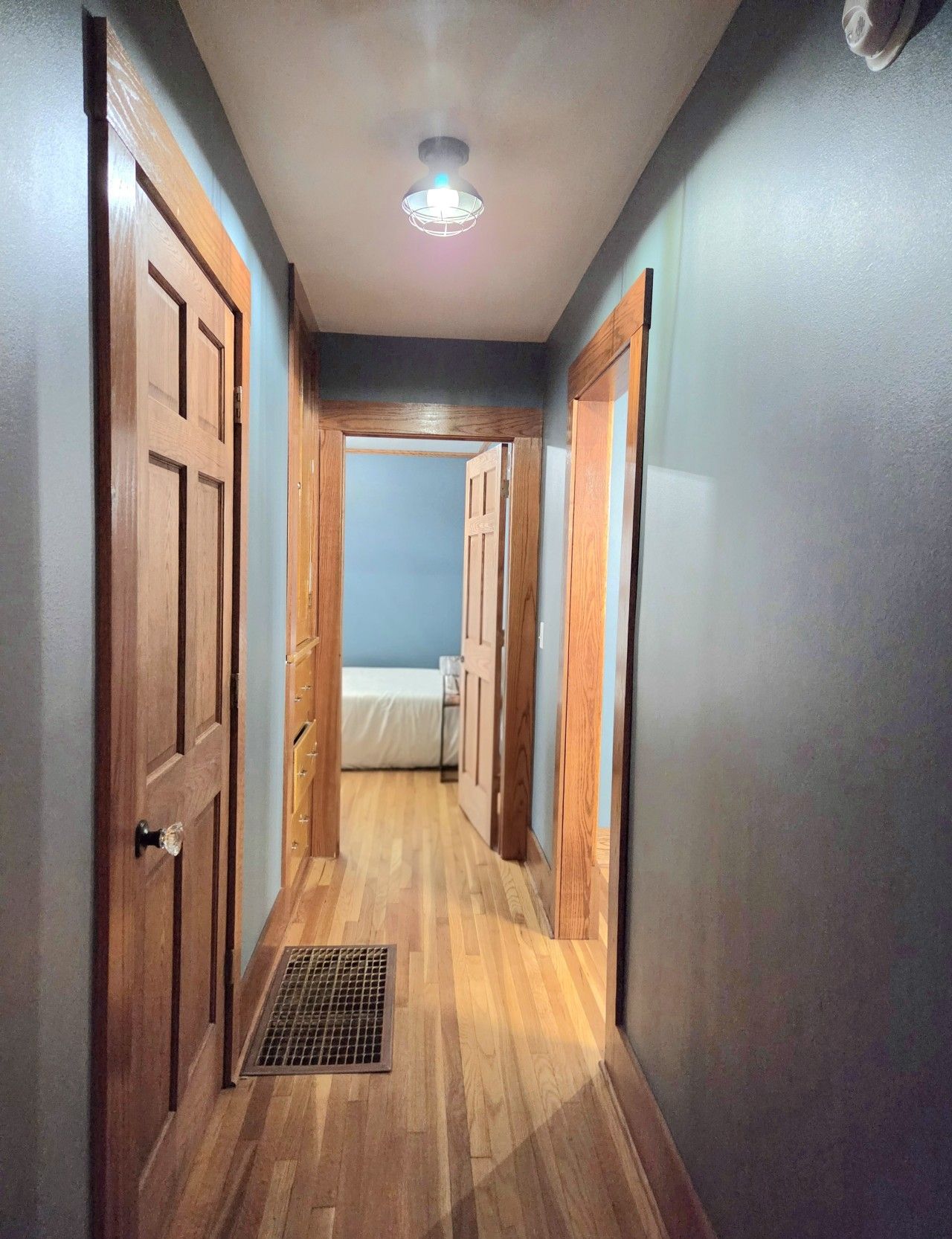 A narrow hallway with wooden doors on both sides, a vent on the floor, and a ceiling light.