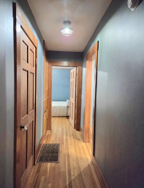 A narrow hallway with wooden doors on both sides, a vent on the floor, and a ceiling light.