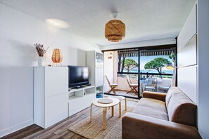 A modern living room with a flat-screen TV, a sofa, a coffee table, wicker pendant lighting, and a view of the outdoors. - Le Cocon Ensoleillé - Avec vue mer (Le Barcarès)