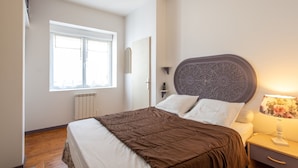 Una camera da letto con un letto, comodino, lampada, finestra e termosifone.