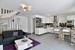 Eine moderne Küche mit weißen Schränken, eine Essecke mit einem weißen Tisch und Stühlen sowie ein Wohnbereich mit einem schwarzen Sofa und einem weißen Couchtisch.