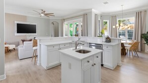 Eine moderne Küche mit weißen Schränken, einer zentralen Kochinsel mit Spüle, einem Deckenventilator und einem großen Fenster mit Vorhängen.