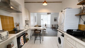 A kitchen with a stove, oven, and washing machine, a dining area with a table and chairs, and a window with blinds. - Charming, spacious and full of natural light 2-story flat in central Saxmundham. (Saxmundham)