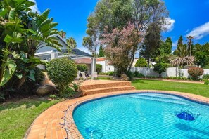 A swimming pool with a tiled deck, surrounded by lush greenery and a white building in the background. - Modern 4BR Sandton Home  Pool, Office, Backup Power & Fast WiFi (Sandton)