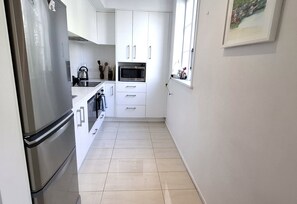 A narrow kitchen with white cabinets, a stainless steel refrigerator, and a microwave. - Coastal Retreat Herne Bay Near Beach and Cafes (Auckland)