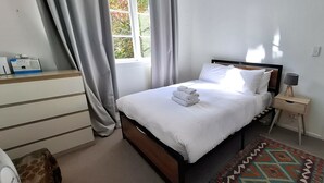 Ein ordentlich bezogenes Bett mit weißen Leinentüchern und einem gefalteten Handtuch, ein Nachttisch mit Lampe, ein gemustertes Teppich und ein Fenster mit Vorhängen.