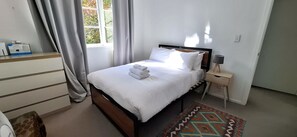 A neatly made bed with white linens and a folded towel, a bedside table with a lamp, a patterned rug, and a window with curtains. - Coastal Retreat Herne Bay Near Beach and Cafes (Auckland)