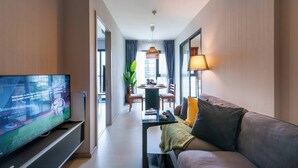 Moderni hotellihuone, jossa on taulutelevisio, sohva, ruokapöytä ja suuri ikkuna.