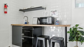 Cocina compacta equipada con microondas, cafetera y pequeño refrigerador. Incluye dos taburetes y una planta en maceta.