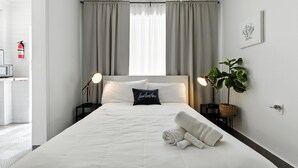 Habitación de hotel con cama, dos lámparas de noche, una planta en maceta, mesita de noche con lámpara, toalla doblada y extintor de pared.