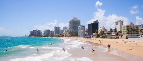 A beach with a city skyline in the background, including high-rise buildings and palm trees.