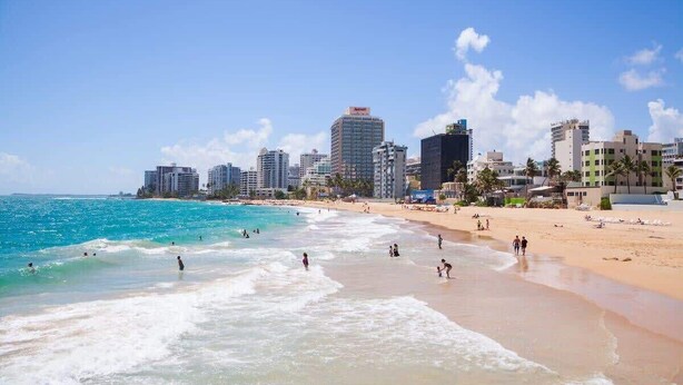 A beach with a city skyline in the background, including high-rise buildings and palm trees. - 1BR 1BA Sleeps 3 Wi-Fi  Walk to Beach (San Juan)