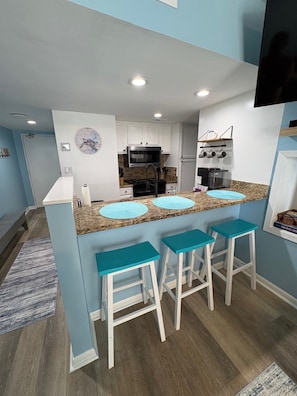 A modern kitchen with white cabinets, a microwave, and a stove. There are three blue bar stools and a granite countertop. - Oceanfront Escape w/ Balcony + 4 pools (Orange Beach)