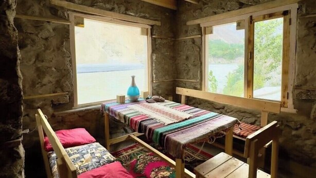 A rustic stone room with a wooden bed, a small table, and two windows. - Hunza Nimbus hotel  (Karimabad)