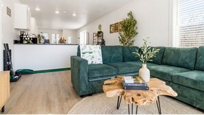 Un salon moderne avec un canapé vert, une table basse et une bibliothèque. On aperçoit à l’arrière-plan un coin cuisine avec des meubles blancs et une plante.
