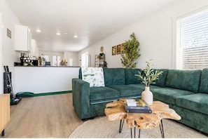 Een moderne woonkamer met een groene bank, een salontafel en een boekenkast. Achtergrond is een keuken met witte kasten en een plant.