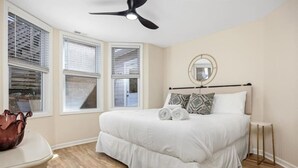 Ein Schlafzimmer mit Bett, Deckenventilator, Nachttisch, Spiegel und einem Fenster mit Jalousien.