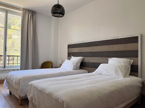 Una habitación con dos camas, una lámpara colgante, una silla y un ventanal con cortinas.